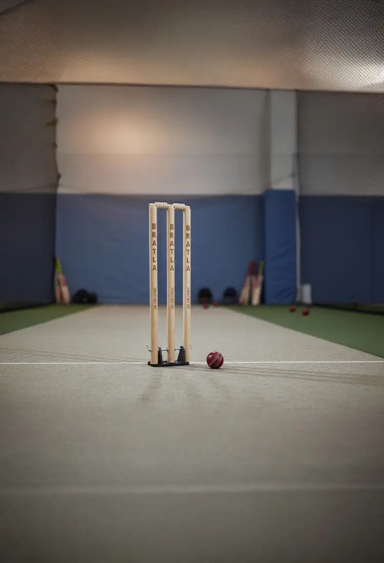 Set of three light-colored wooden cricket stumps with “bratla” branding, standing upright on a pitch beside a red leather cricket ball.